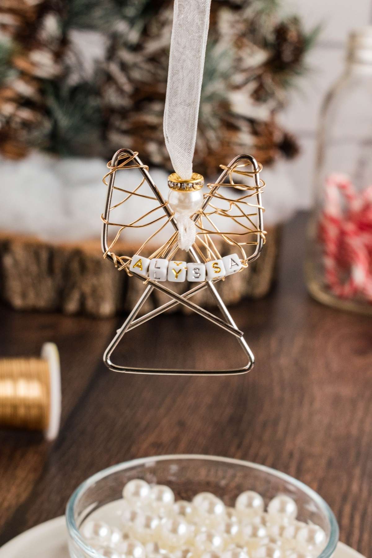 Wire angel ornament with the name "Alyssa" spelled in white beads, hanging from a ribbon. On the table are pearls, a spool, and blurred festive decor in the background.