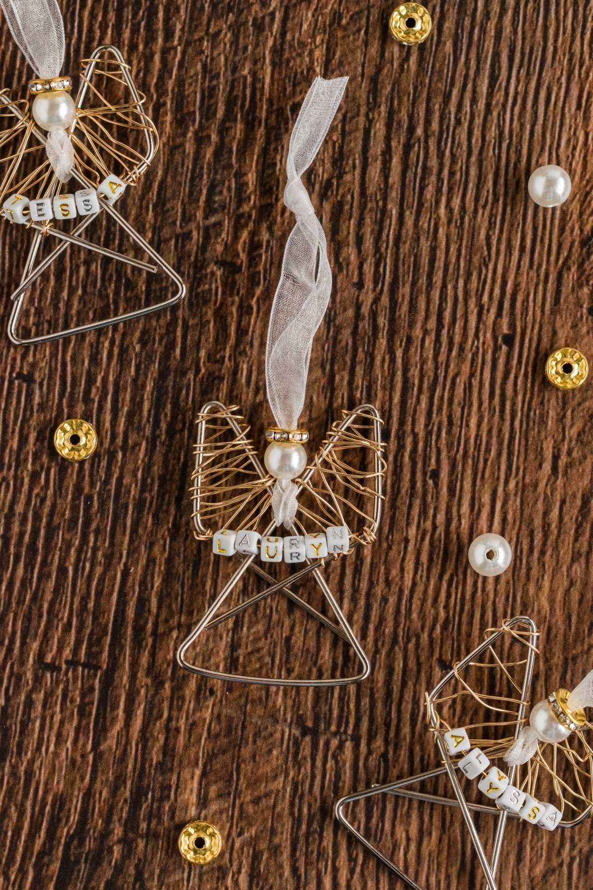 Wire angel ornaments with beaded names are displayed on a wooden surface, adorned with golden sequins and white pearls.