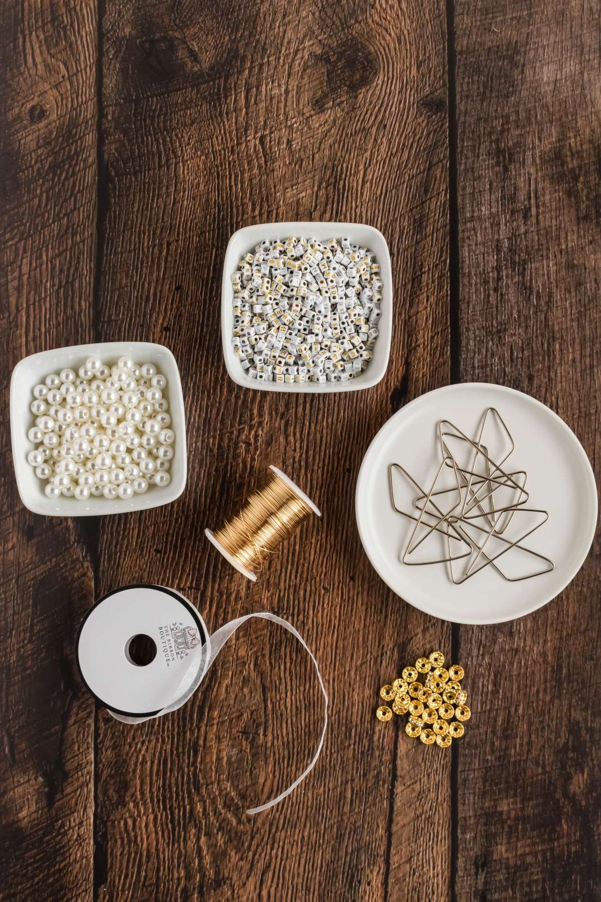Assorted jewelry-making supplies on wooden surface: white pearls, gold beads, metallic stars, spool of gold wire, and ribbon spool.