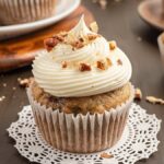 A hummingbird cupcake with cream cheese frosting and chopped nuts sits elegantly on a doily, surrounded by more cupcakes and a rustic wooden board backdrop.