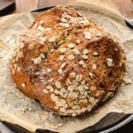 A loaf of round Irish soda bread topped with oats, resting on parchment paper in a black pan.