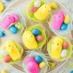 Yellow and pink marshmallow chicks nestle in Easter Peep Cupcakes adorned with colorful speckled eggs, all elegantly arranged on a plate. Around it, scattered eggs and shredded paper create a festive display on the white surface.