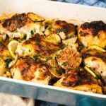 Baked chicken thighs in a dish with lemon slices, roasted garlic cloves, herbs, and capers, reminiscent of the classic Chicken with 20 Cloves of Garlic dish.