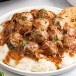 A plate of slow cooker French onion meatballs nestles atop mashed potatoes, crowned with caramelized onions and garnished with parsley, perfectly accompanied by slices of bread.
