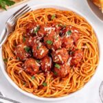 A bowl of spaghetti features slow cooker meatballs in tomato sauce, topped with grated cheese and herbs. A fork rests on the plate's edge alongside a side of garlic bread and a small bowl of red pepper flakes nearby.