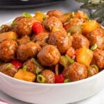 A white bowl filled with Slow Cooker Sweet and Sour Meatballs, pineapple, and colorful bell pepper pieces, garnished with chopped green onions. A red and white striped cloth is partially visible nearby.