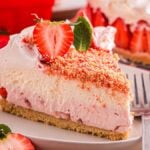 A slice of strawberry shortcake cheesecake topped with fresh strawberries and crumb topping sits elegantly on a white plate, accompanied by a shining fork.