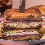 A Rachel sandwich, stacked with layers of savory meat, cheese, and tangy coleslaw, is perfectly sliced by a knife on a plate, accompanied by crispy chips and a crunchy pickle slice.