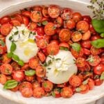 Oval dish featuring a delightful tomato burrata salad with sliced cherry tomatoes, two creamy balls of burrata cheese, fresh basil, and thyme sprigs.