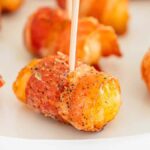 Bacon-wrapped tater tots, secured with toothpicks and seasoned with a dash of pepper, are artfully arranged on a white plate.
