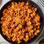 A skillet filled with delicious Mediterranean chicken pasta, featuring spiral noodles, tender chicken, and vibrant vegetables in a red sauce. Nearby, a striped cloth, lemon wedges, olives, and parsley complete the enticing presentation.