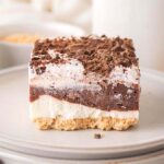 A layered chocolate delight with a crumbly base and creamy layers under a rich chocolate topping, elegantly stacked on gray plates. Assorted ingredients are artistically scattered in the background.
