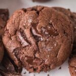 Close-up of multiple chocolate fudge cookies with rich chocolate chunks, surrounded by pieces of decadent chocolate, on a light surface.