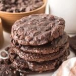 A tempting stack of flourless fudge cookies adorned with chocolate chips, surrounded by a bowl of chocolate chips, cocoa powder, and a refreshing glass of milk.