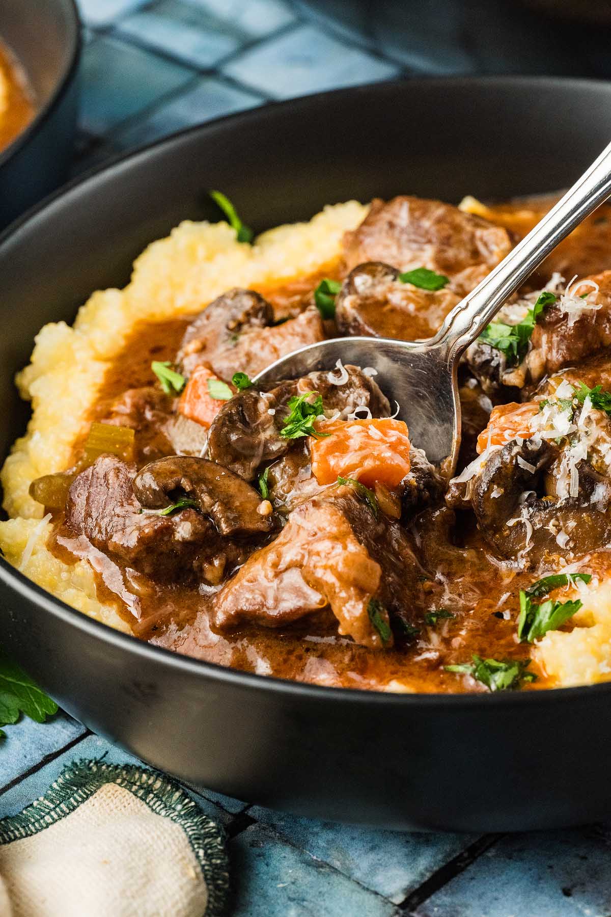 A bowl of creamy polenta topped with Italian beef stew, carrots, mushrooms, and fresh herbs, served with a spoon.