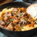 A bowl of Italian beef stew with carrots and herbs served over polenta, accompanied by a slice of bread.