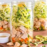 Three mason jars filled with a delightful Mason Jar Chicken Zoodle Soup, featuring spiralized zucchini, tender chicken pieces, and diced vegetables on a wooden board. A small dish of dressing accompanies this fresh and savory treat.
