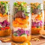 Four mason jars brimming with vibrant Asian chicken salad ingredients, including crisp lettuce and sliced vegetables, rest on a wooden board. The colorful layers promise a delightful crunch, accompanied by a small bowl for extra dressing or toppings.
