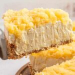 A slice of creamy, no-bake pineapple cheesecake on a spatula above the rest of the cake, featuring a crumbly crust and pineapple topping. Plates and utensils are blurred in the background.