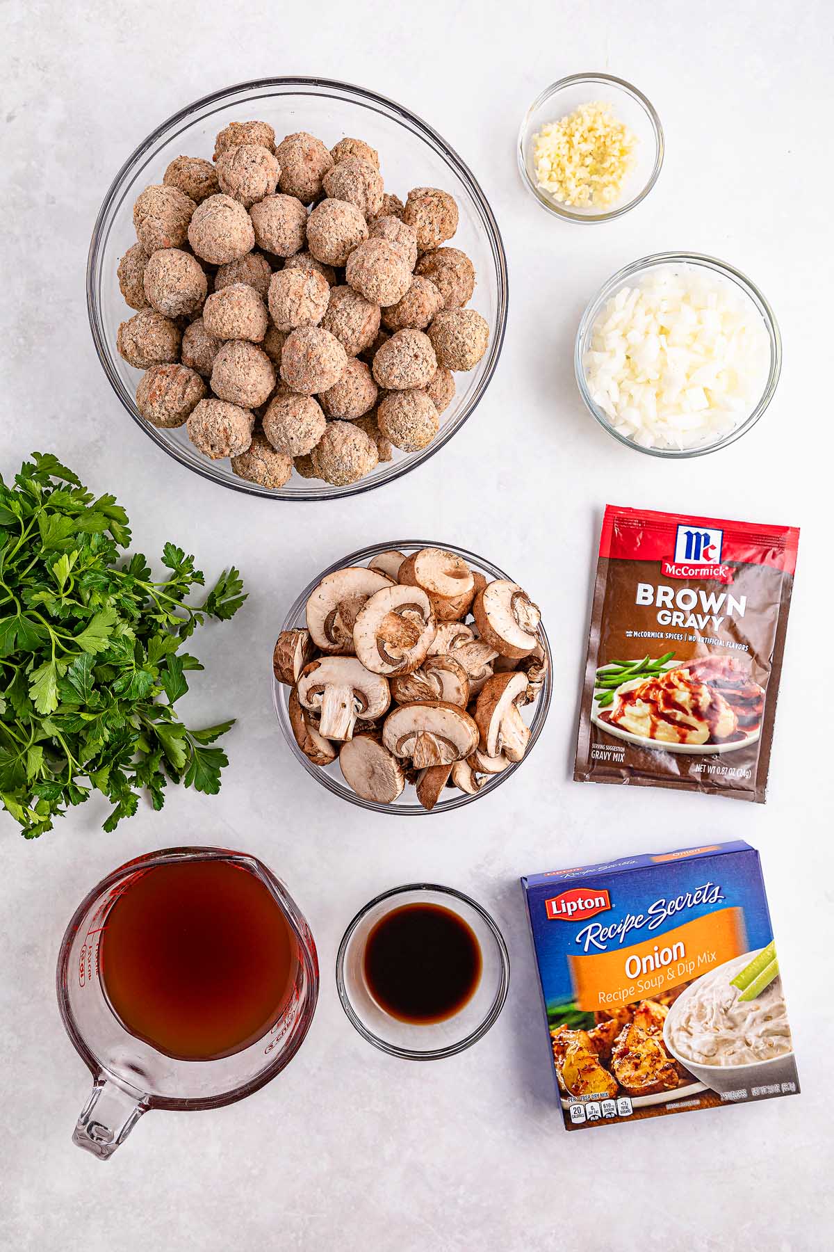 Ingredients on a counter: raw meatballs, minced garlic, chopped onions, and sliced mushrooms ready to transform into delicious slow cooker Salisbury steak. Add a brown gravy packet, beef broth, Worcestershire sauce, onion soup mix, and fresh parsley for the perfect blend of flavors.