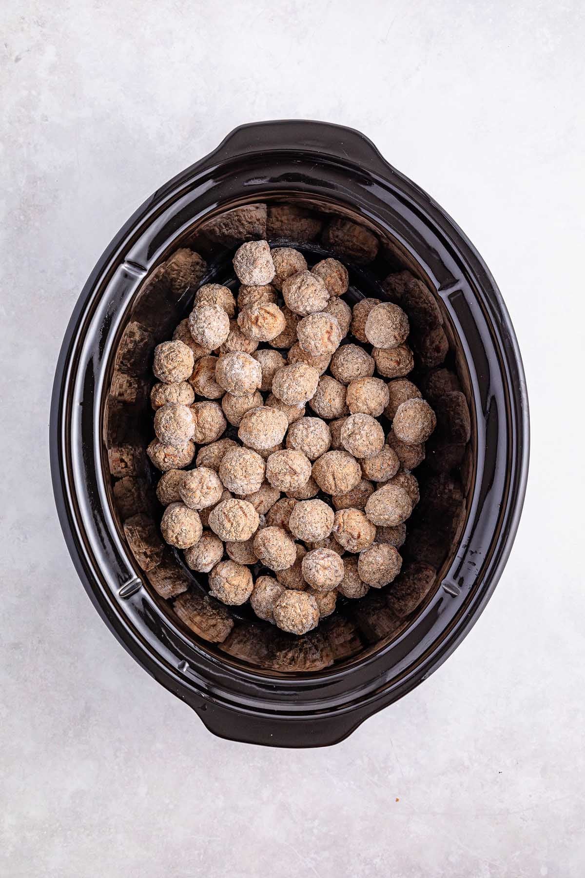 A slow cooker brimming with uncooked, seasoned Salisbury steak meatballs rests on a light background.
