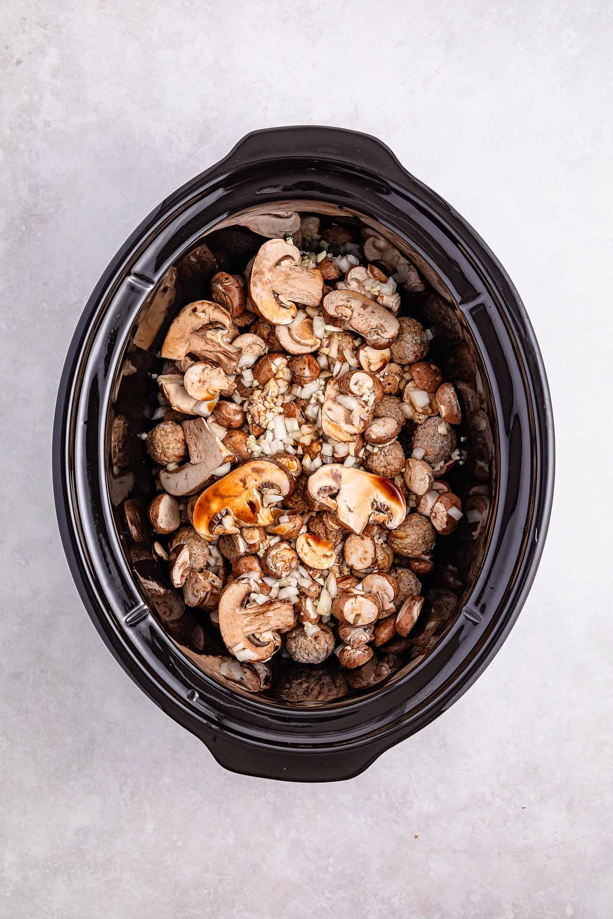 A slow cooker brimming with sliced mushrooms, onions, and whole potatoes on a light surface offers a cozy base for mouthwatering Salisbury steak meatballs.