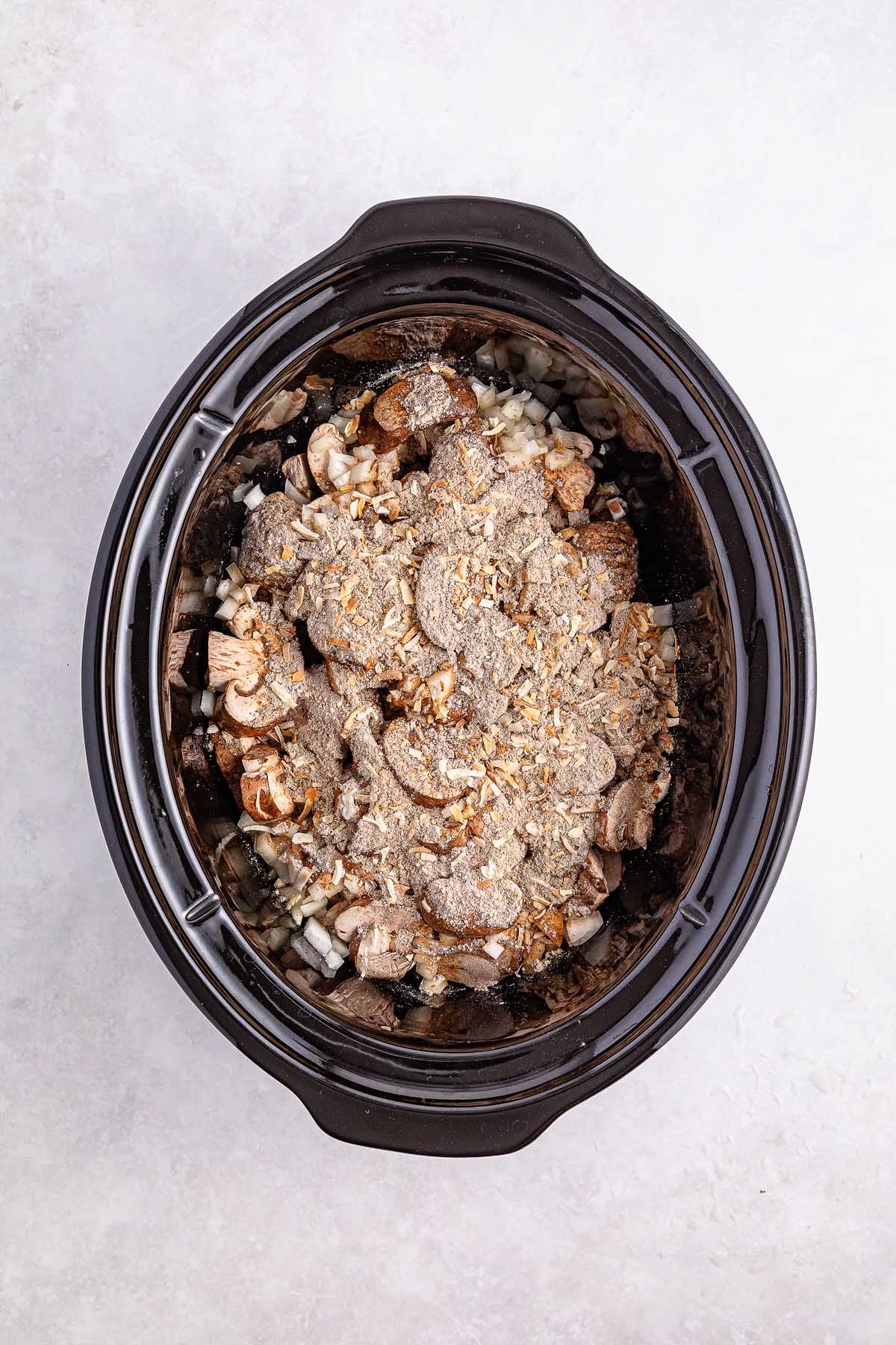 The slow cooker is filled with savory Salisbury steak meatballs, joined by sliced mushrooms, chopped onions, and a flavorful seasoning mix on a light surface.