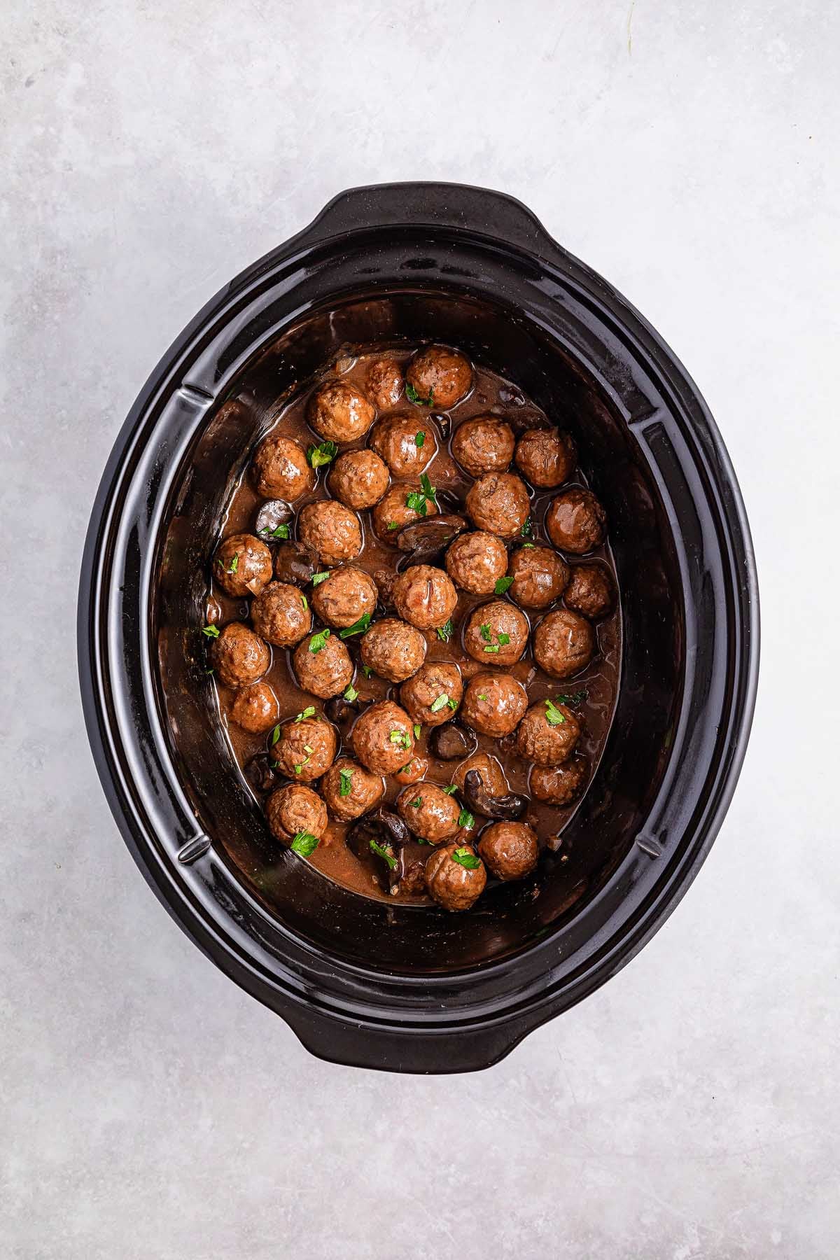 Salisbury steak meatballs simmer in a rich brown sauce inside a black slow cooker, garnished with chopped herbs.