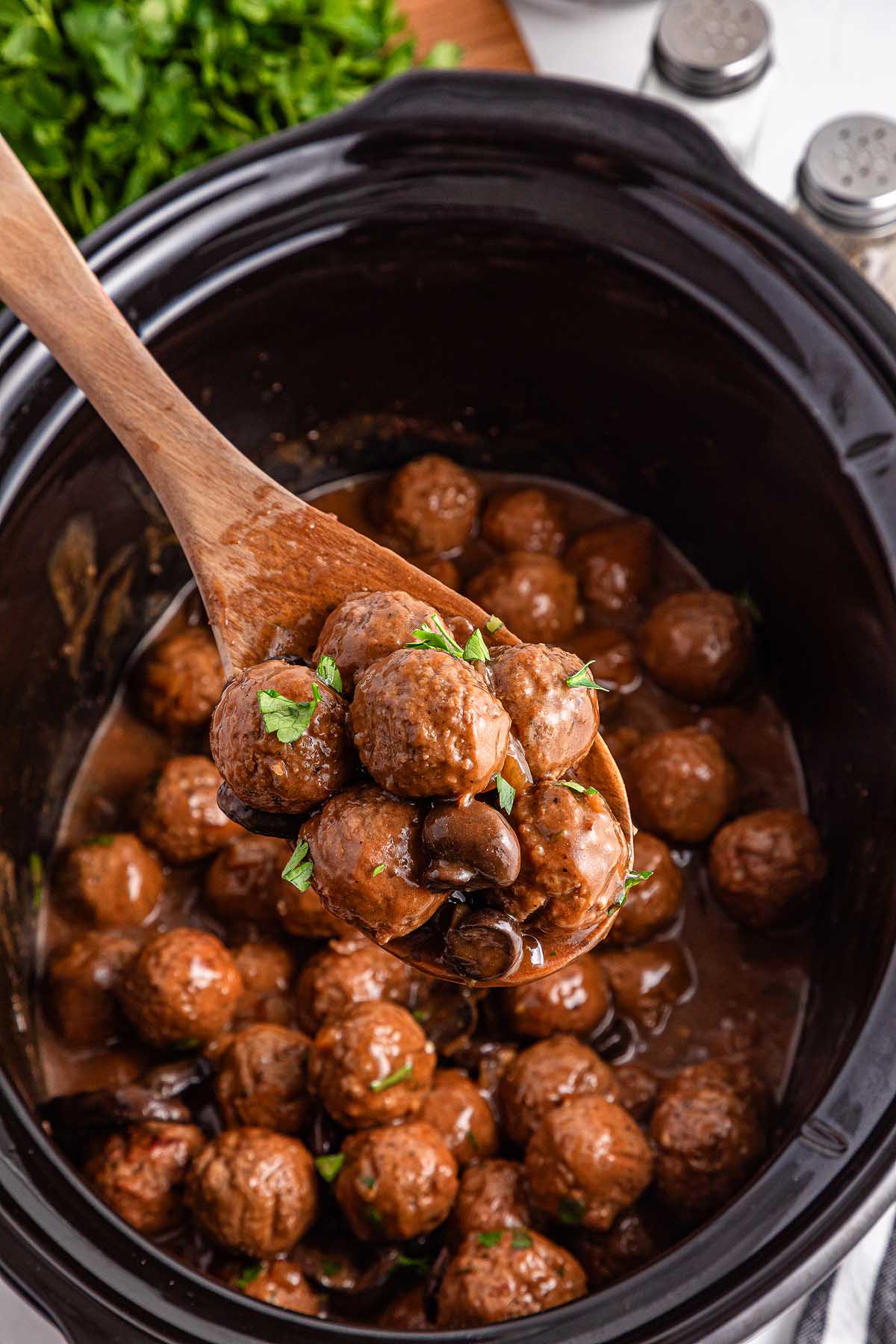A wooden spoon holds savory Salisbury steak meatballs over a slow cooker filled with more meatballs in a rich brown sauce, garnished with chopped parsley.
