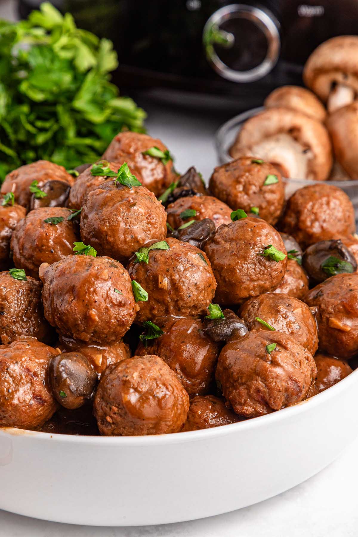 A bowl of saucy Salisbury steak meatballs garnished with herbs, evoking the cozy comfort of a slow cooker. Mushrooms and a bunch of greens linger invitingly in the background.