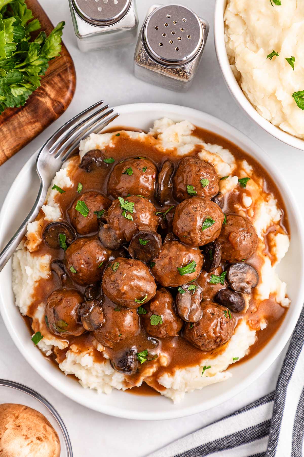 Delicious slow cooker Salisbury steak meatballs and mushrooms nestled in gravy over creamy mashed potatoes, garnished with fresh parsley. A fork waits on the plate, with salt and pepper shakers nearby to complete this comforting meal.