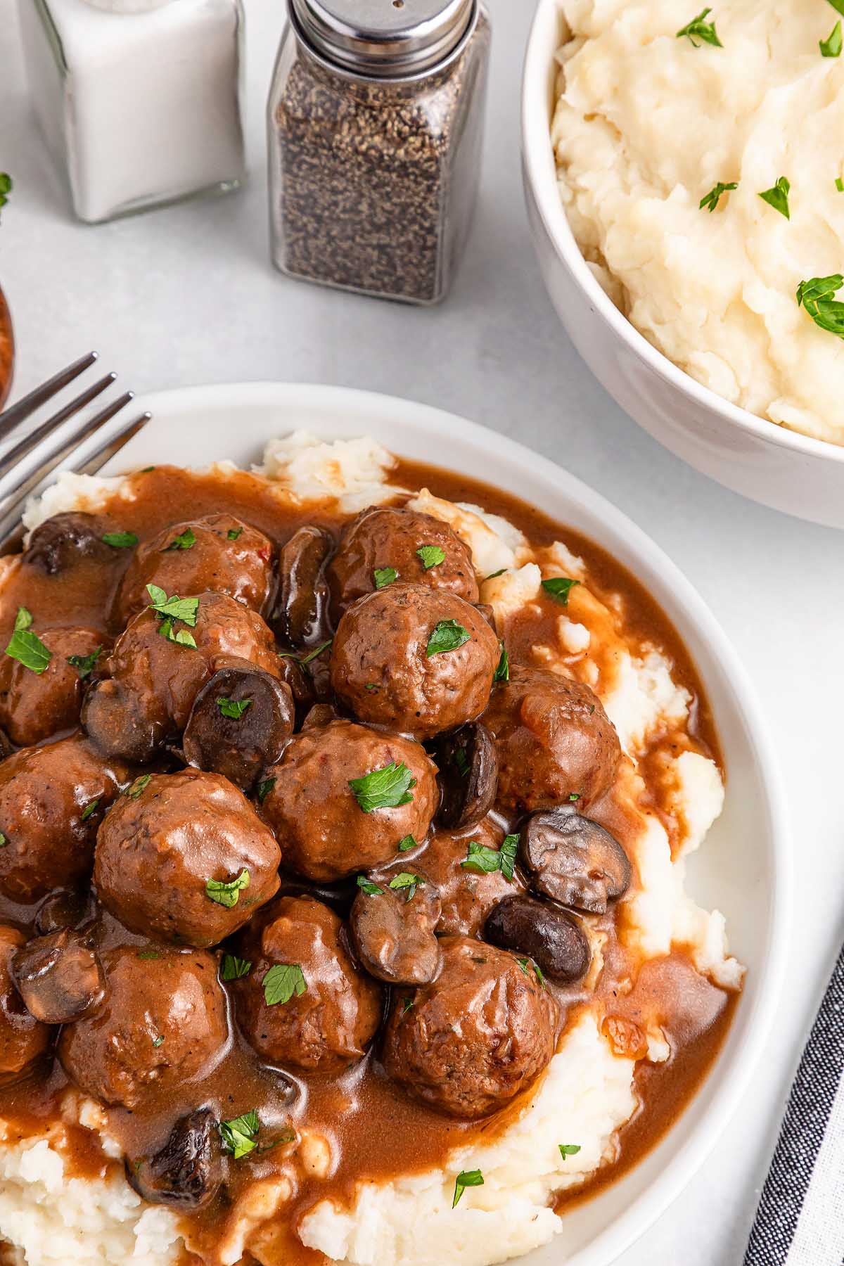 Slow cooker Salisbury steak meatballs with gravy and mushrooms are served over creamy mashed potatoes, garnished with parsley. A bowl of mashed potatoes and salt and pepper shakers add a cozy touch to the background.