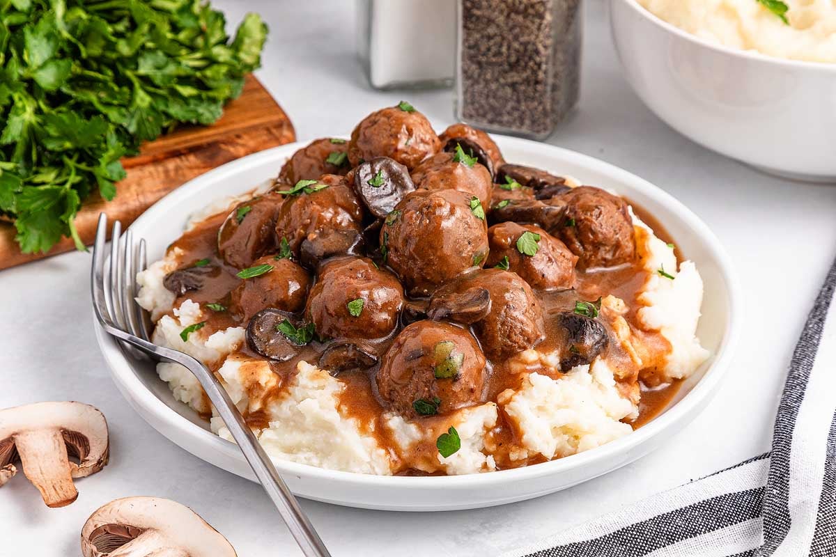 A plate of mashed potatoes topped with savory slow cooker Salisbury steak meatballs and rich mushroom gravy, garnished with fresh parsley. A fork rests beside the dish, while a sprinkle of parsley and seasonings sets the scene in the background.