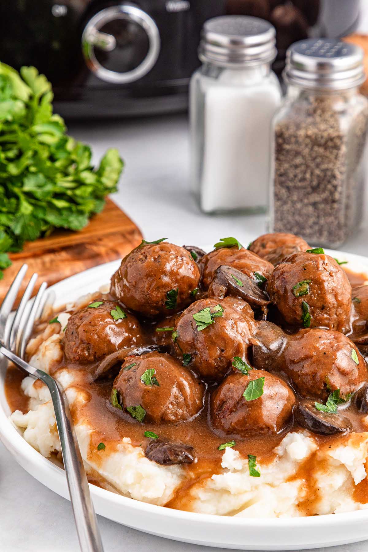 A delicious plate of slow cooker Salisbury steak meatballs smothered in rich gravy over creamy mashed potatoes, garnished with fresh parsley. A fork sits invitingly on the side, with salt and pepper shakers and vibrant parsley adding a touch of freshness to the background.