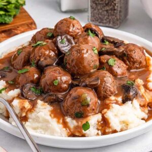 A plate of slow cooker Salisbury steak meatballs and mushrooms in brown gravy served over mashed potatoes, garnished with parsley. A fork rests on the plate.