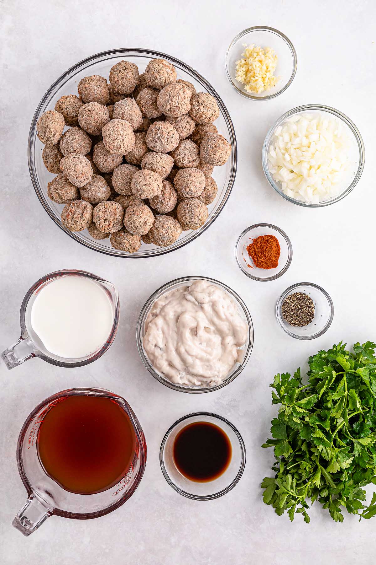 Ingredients on a table: a bowl of meatballs ready for the slow cooker, chopped onions, minced garlic, chopped parsley, pepper, paprika, two measuring cups with broth and milk, a bowl of sauce for Swedish meatballs, and a small dish of soy sauce.
