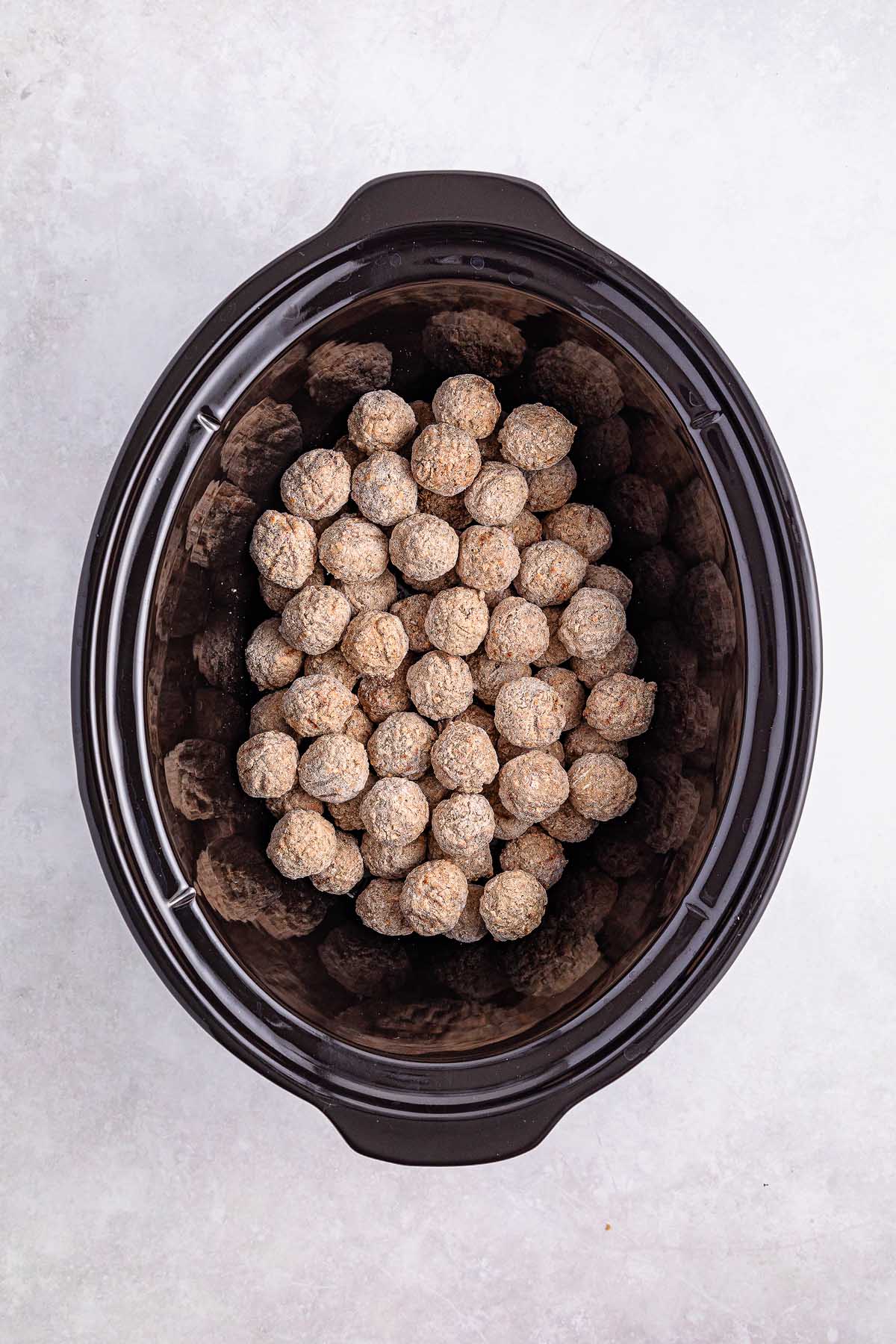 A slow cooker brimming with uncooked Swedish meatballs rests on a gray surface.