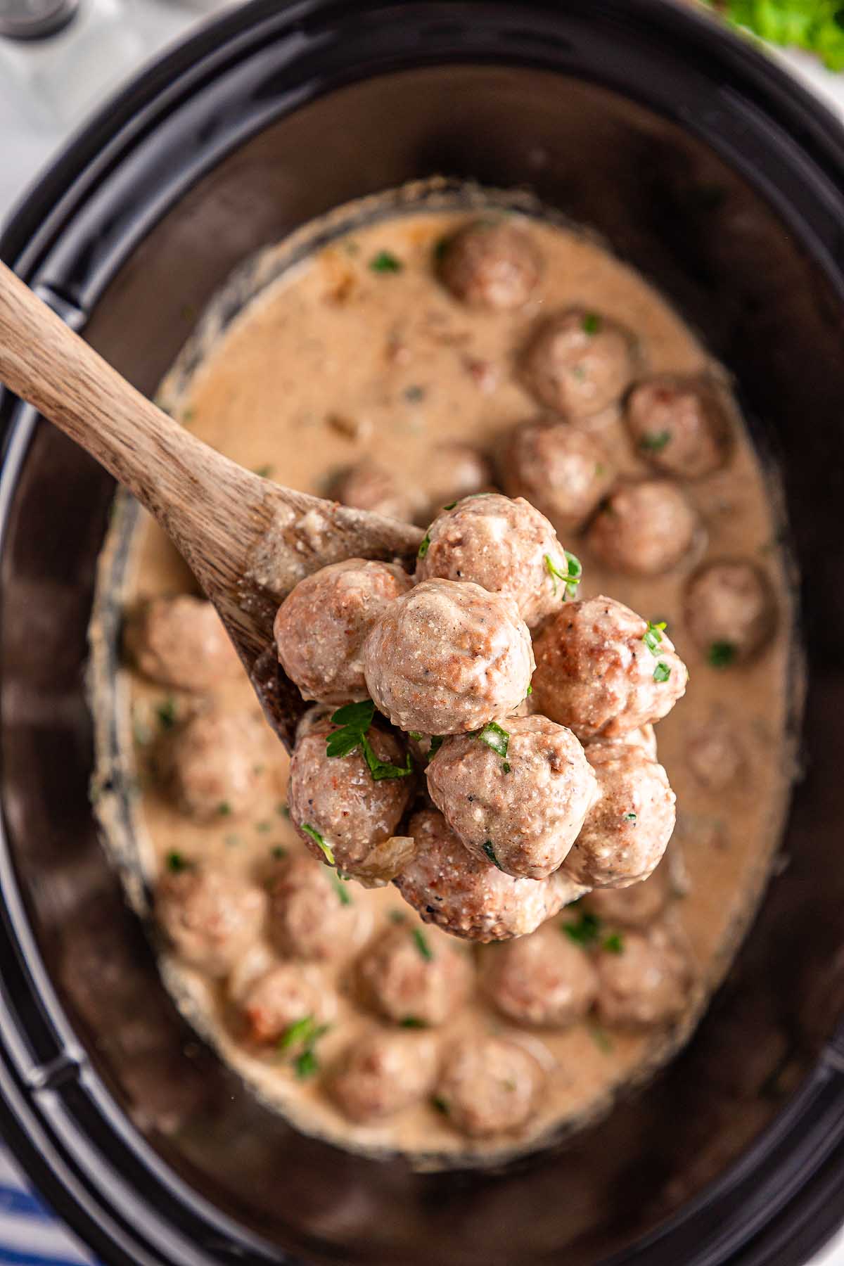A wooden spoon gently cradles a batch of slow cooker Swedish meatballs, dripping with a creamy sauce and topped with a sprinkle of fresh parsley.