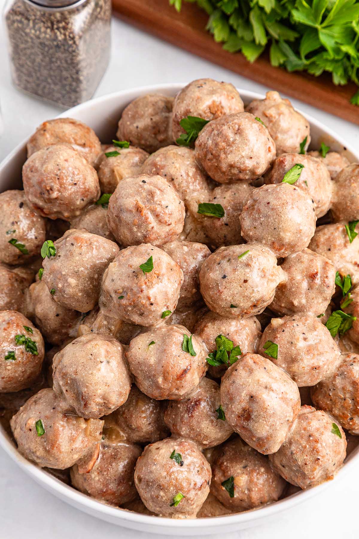 A bowl filled with creamy slow cooker Swedish meatballs, garnished with chopped parsley. A pepper shaker and fresh parsley are in the background, completing the cozy setup.