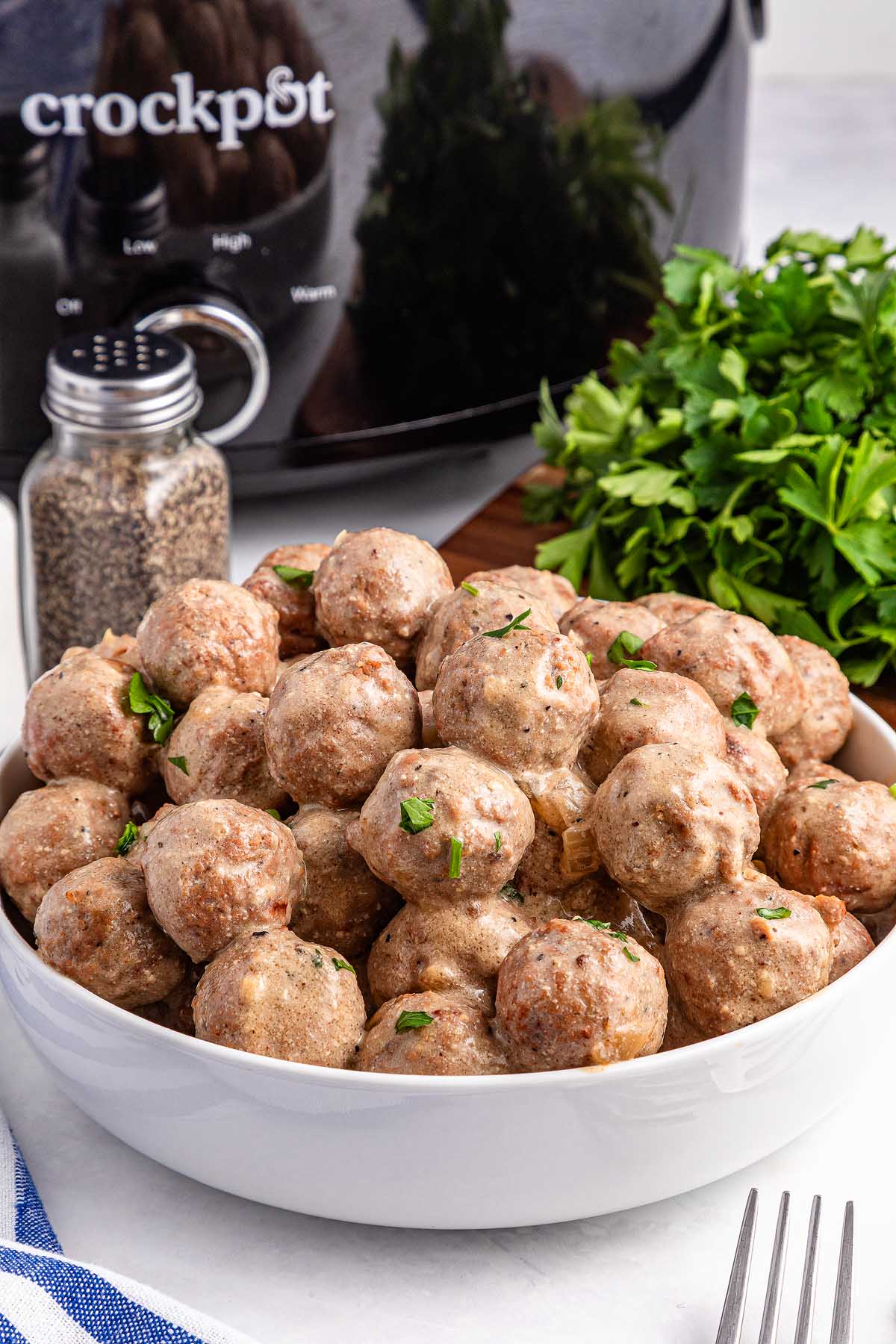A bowl of delicious Swedish meatballs garnished with herbs sits enticingly in front of a slow cooker. A pepper shaker and fresh parsley add the perfect finishing touches.