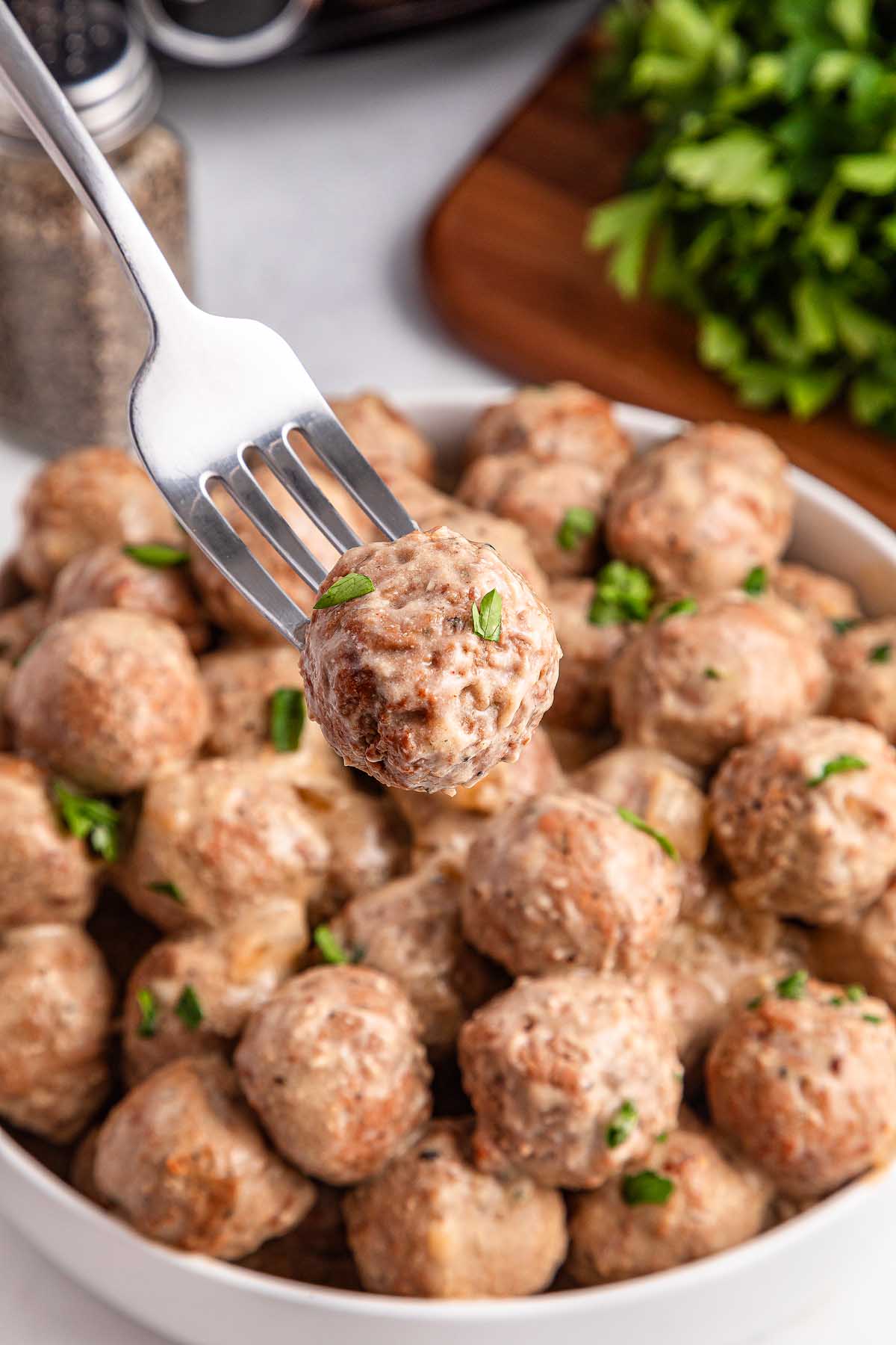 A fork holding a savory meatball over a bowl of Crockpot Swedish meatballs, garnished with fresh green herbs.