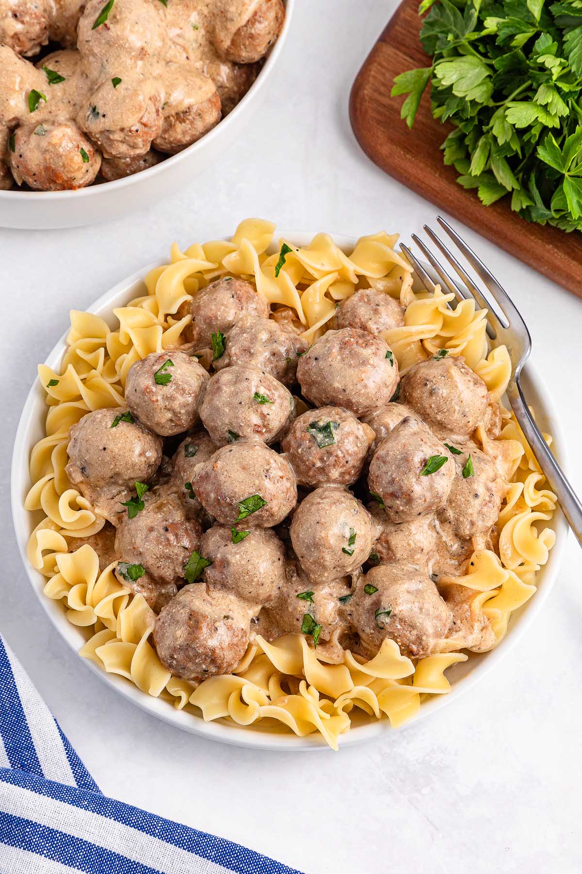 A plate of pasta topped with slow cooker Swedish meatballs in a creamy sauce, garnished with chopped parsley. A fork rests on the side, and sprigs of fresh parsley are visible in the background.