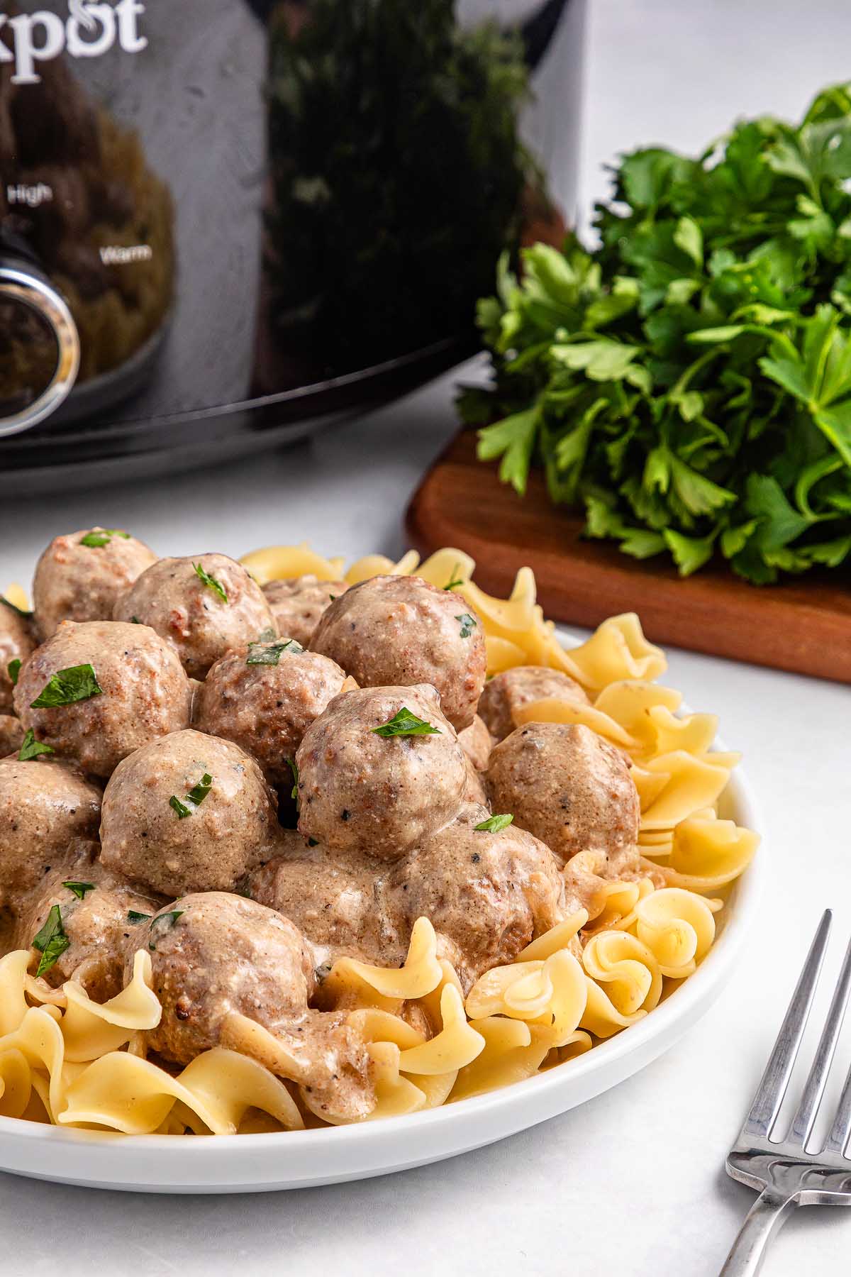 A plate of slow cooker Swedish meatballs in creamy sauce served over egg noodles, with a parsley garnish. A bunch of fresh parsley and the trusty slow cooker linger in the background, setting a cozy culinary scene.