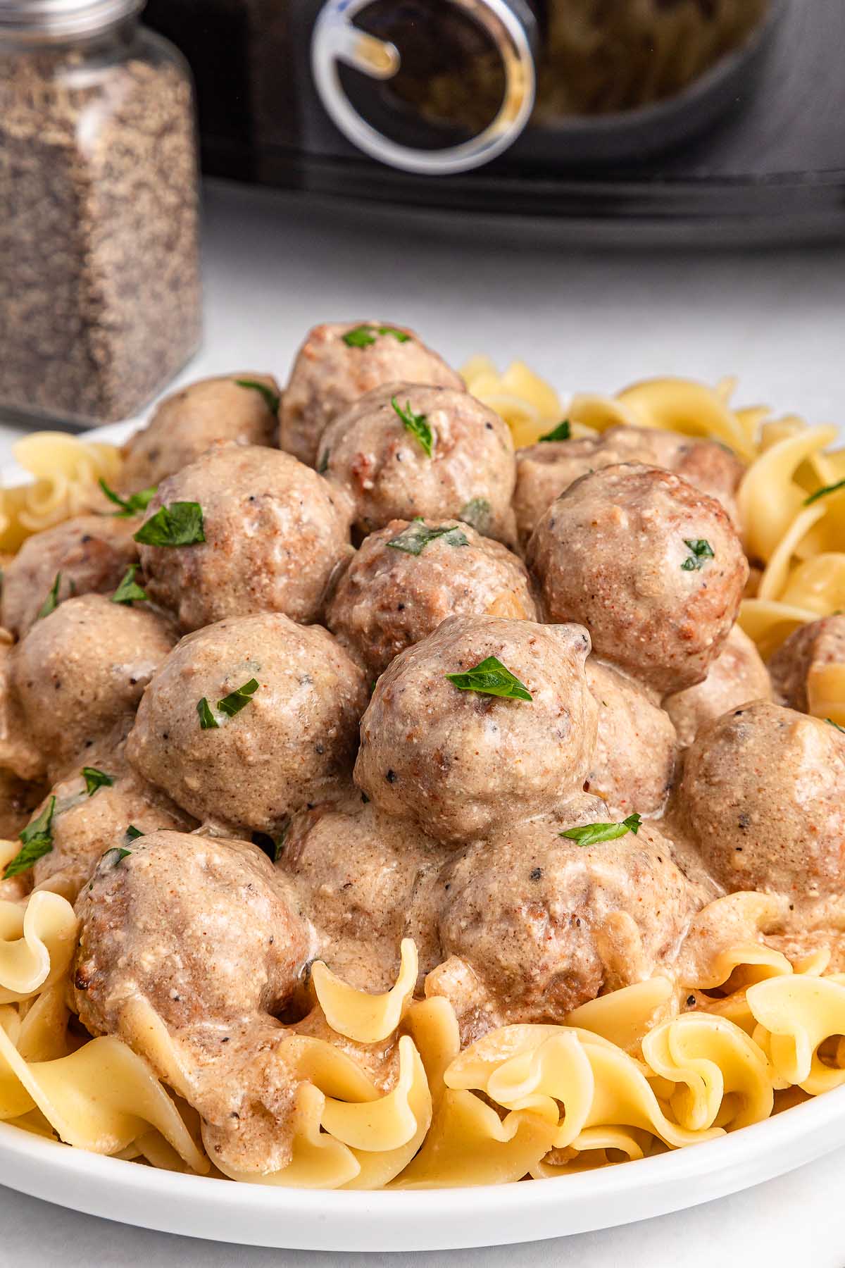 A plate of creamy Swedish meatballs topped with chopped herbs is elegantly served over a bed of cooked egg noodles. In the background, a container of black pepper and a trusty slow cooker hint at the secret behind this delightful dish.