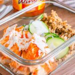 Buffalo chicken meal prep bowl featuring a glass container filled with savory buffalo chicken, crisp sliced cucumbers, and fluffy rice drizzled with dressing. A napkin, fork, and knife are neatly positioned beside it while a jar of hot sauce sits in the background.