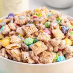 A bowl of colorful bunny bait snack mix with cereal, candies, and sprinkles sits on a table alongside a bunny-themed napkin.