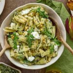 A bowl of cavatelli with pesto, ricotta, arugula, and pine nuts is artfully surrounded by small dishes and flowers.