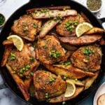 Skillet with seasoned Chicken Vesuvio-style thighs, potato wedges, and peas, garnished with lemon slices and herbs. Nearby are parsley, pepper, a bowl of lemon wedges, wine, and a plate with bread slices.