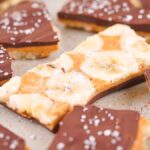 On a baking sheet, chocolate and banana bark pieces are elegantly topped with sea salt, creamy peanut butter, and luscious white chocolate.