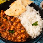 A delightful chickpea curry sits in a bowl alongside fluffy white rice, topped with naan pieces and garnished with cilantro. Spices, ginger, lime, and a cloth adorn the tiled surface nearby.
